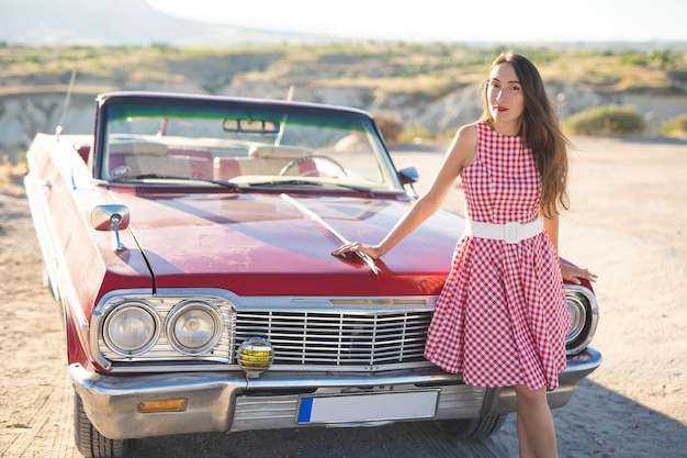 Foto hermosa chica en un vestido retro al amanecer feliz posando en el fondo del paisaje de la montaña en capadocia cerca de un coche retro rojo