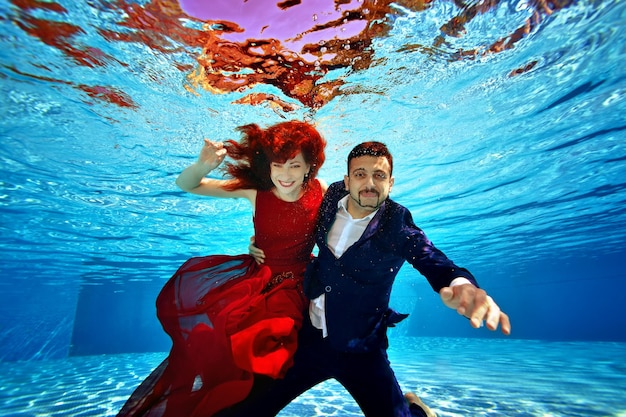 Una hermosa chica con un vestido rojo y un chico con traje están nadando bajo el agua en la piscina