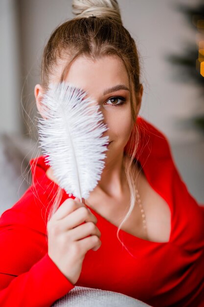 Una hermosa chica con un vestido rojo posa en el interior de una casa sentada en un sofá cubre la mitad de su cara con un bolígrafo y mira a la cámara