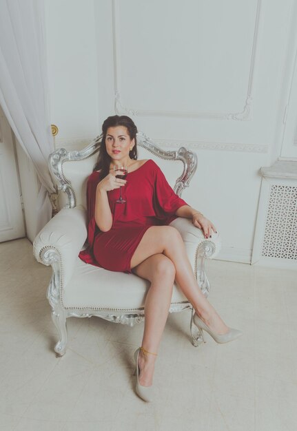 Hermosa chica con vestido rojo posando sentada en un sillón vintage con una copa de vino.