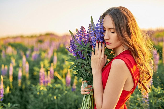 Hermosa chica con un vestido rojo con un ramo de flores de lupino en el campo