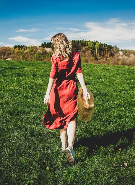 Hermosa chica en vestido rojo y sombrero tiene un tiempo sin preocupaciones en la pradera en las montañas por un bosque. Temporada de primavera