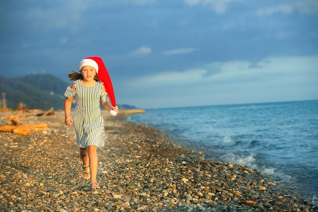 una hermosa chica con un vestido y un sombrero rojo de santa claus corre alegremente a lo largo de la orilla del mar