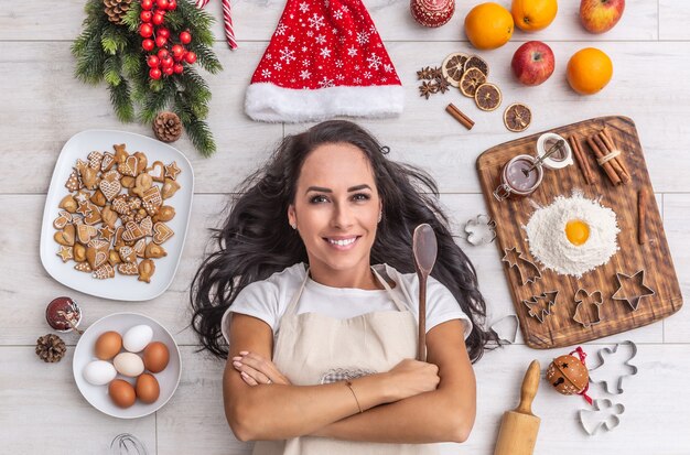 Hermosa cocinera de pelo oscuro tendida y sonriendo ampliamente en el suelo, sosteniendo la cuchara de madera y rodeada de pan de jengibre, huevos, harina, sombrero de navidad, naranjas secas y formas para hornear.