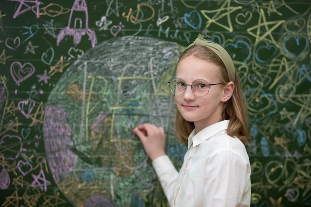 Una hermosa colegiala con anteojos se para en una pizarra pintada y mira a la cámara.