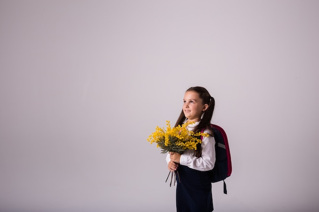 Una hermosa colegiala morena en uniforme con una mochila se encuentra con un ramo de mimosa sobre un fondo blanco con un lugar para el texto