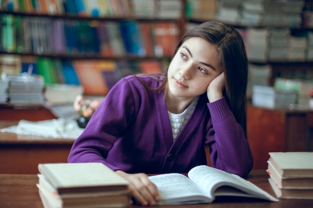 Hermosa colegiala sentada en la biblioteca y leyendo un libro