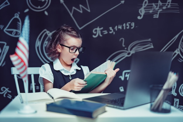 Hermosa colegiala sentada en el escritorio y estudiar en línea con la computadora portátil sobre fondo negro con ...