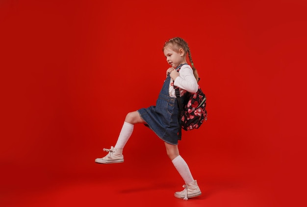 Una hermosa colegiala con un vestido de mezclilla con una mochila sobre los hombros camina sobre un fondo rojo aislado.