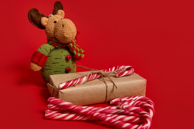Hermosa composición minimalista de artículos navideños, caja de regalo en papel kraft con lazo atado, piruletas dulces y muñeco de peluche de ciervo sobre fondo rojo con espacio para copiar anuncios