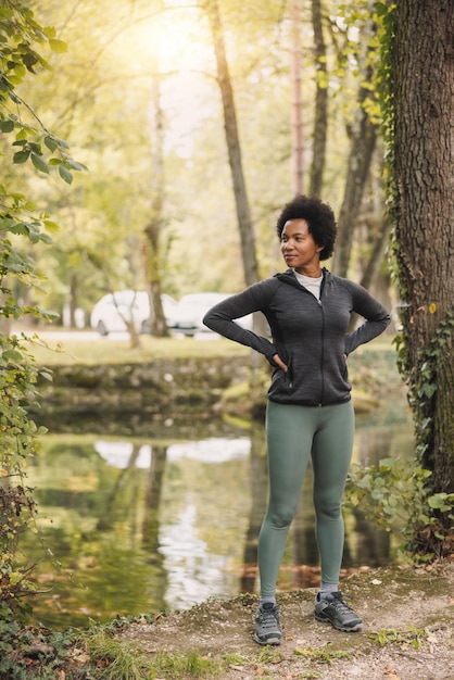 Hermosa corredora afroamericana madura que se relaja después del entrenamiento deportivo por la mañana cerca del río en la naturaleza.