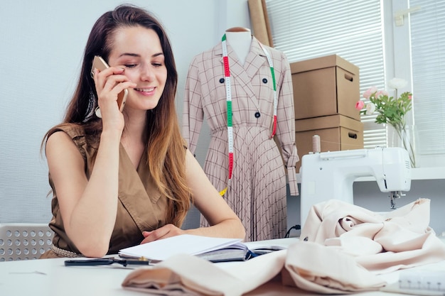 hermosa costurera con cabello largo escribe en un cuaderno. el sastre crea una colección de atuendos y habla por teléfono con el cliente. ropa de diseñador de mujer joven notas ideas en cuaderno