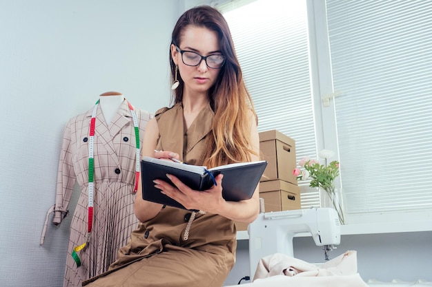 Hermosa costurera con cabello largo escribe en un cuaderno. el sastre crea una colección de trajes. ropa de diseñador de mujer joven notas ideas en cuaderno