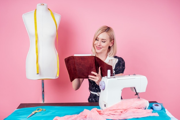 Hermosa costurera rubia escribe en un cuaderno. el sastre crea una colección de trajes cose ropa a máquina de coser en el taller. mujer joven diseñador ropa notas ideas cuaderno rosa studio