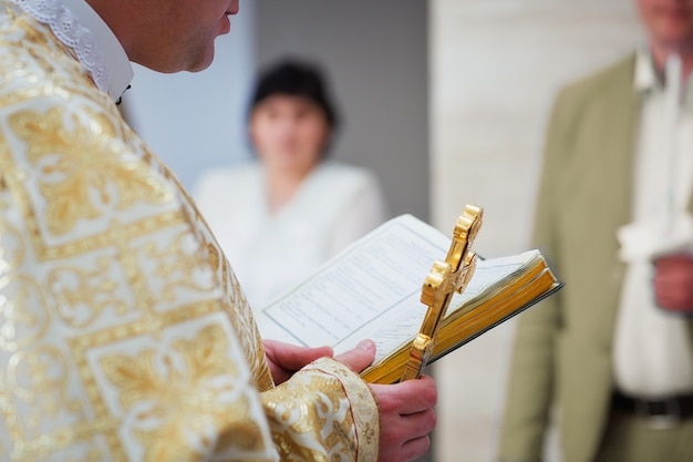 Hermosa cruz de oro en manos masculinas del sacerdote con túnica de oro en ceremonia en la iglesia catedral cristiana, evento sacramental santo. Sacerdote sosteniendo una Biblia