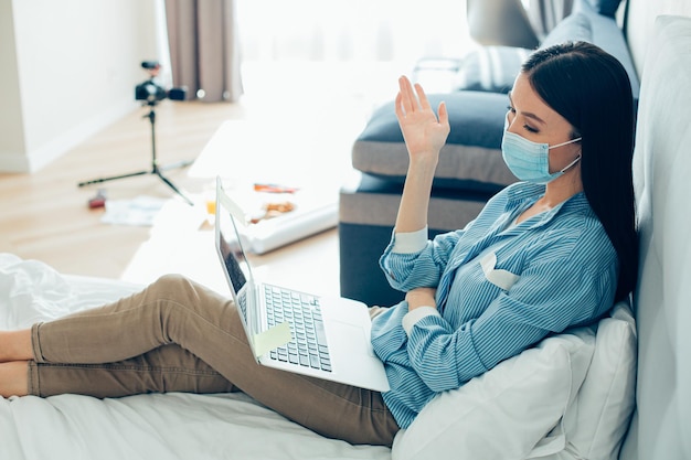 Hermosa dama de cabello largo con una máscara médica sentada en la cama con una computadora portátil en el regazo y mirando la cámara web