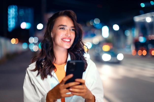 Hermosa dama caminando por la calle de la ciudad y usando un teléfono inteligente en la noche de pie cerca de la carretera libre