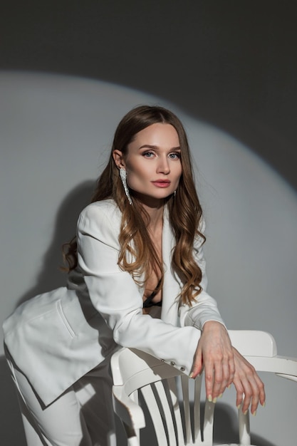 Hermosa dama de moda en un elegante traje blanco se encuentra cerca de una silla blanca sobre un fondo oscuro en el estudio en una fiesta