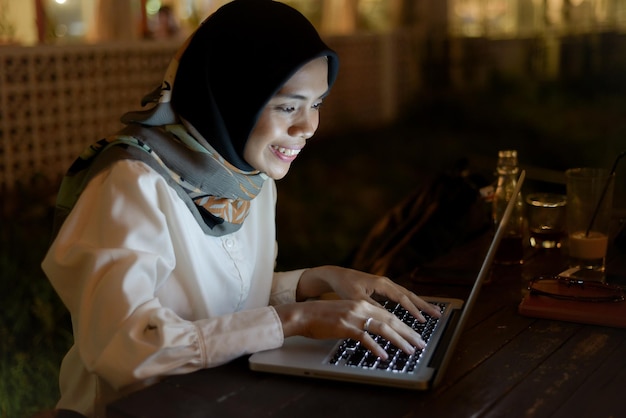 hermosa dama musulmana asiática ropa casual trabajando usando una computadora portátil por la noche en un café al aire libre