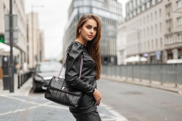 Hermosa dama de negocios con estilo en ropa negra de moda con un vestido y una bolsa de cuero negro de moda está caminando en la ciudad Chica elegante urbana