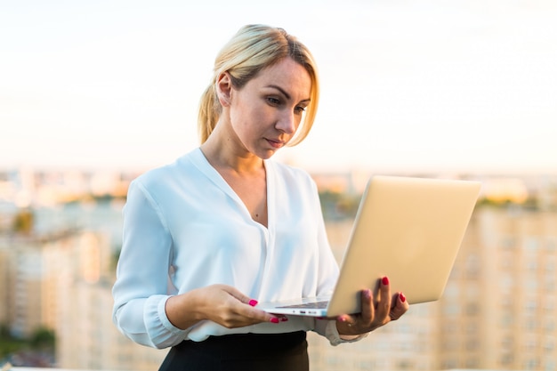 Hermosa dama de negocios inteligente de pie en el techo con la computadora portátil en las manos