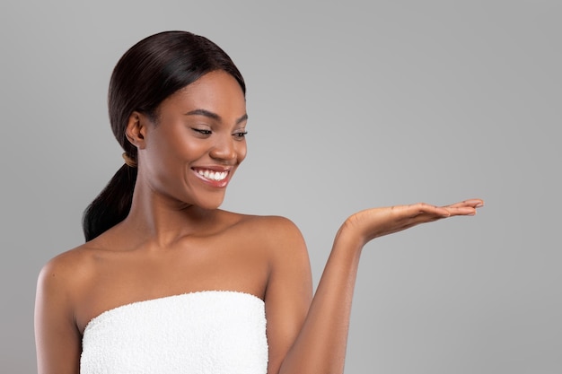 Hermosa dama negra sonriente en una toalla mirando a la mano con un espacio en blanco aislado en el fondo gris