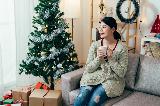 la hermosa dama que sostiene té caliente disfruta de la comodidad del hogar y la alegría de la temporada festiva. una dama sonriente que mira hacia otro lado está descansando en una habitación con adornos navideños. estilo de vida autentico