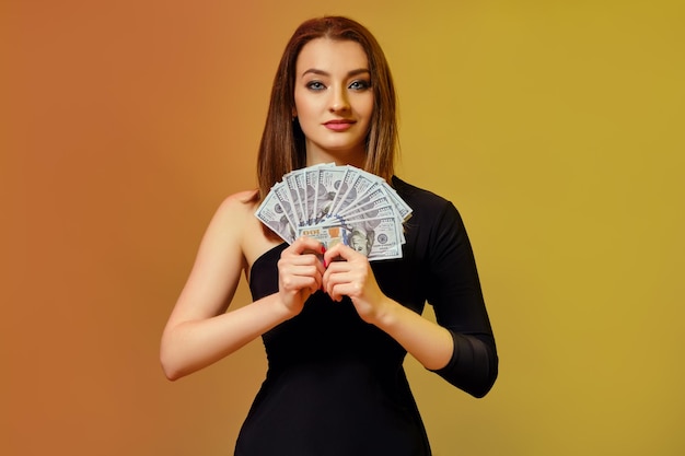 Hermosa dama rubia con maquillaje brillante manicura roja en vestido negro está sonriendo mostrando abanico de billetes de cien dólares posando sobre fondo colorido Casino de póquer de juego Primer plano