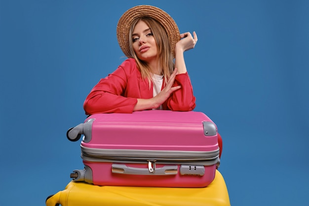 Hermosa dama rubia con sombrero de paja blusa blanca chaqueta roja Ella te está mirando mientras está sentada apoyada en maletas coloridas posando sobre fondo azul Concepto de viaje Espacio de copia de primer plano