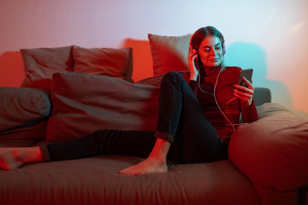 Hermosa dama sentada en el sofá con auriculares y teléfono celular en las manos Mujer joven escuchando música en auriculares mientras pasa tiempo en casa