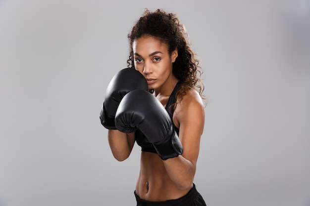 Hermosa deportista africana aislada sobre fondo gris, haciendo ejercicios con guantes de boxeo