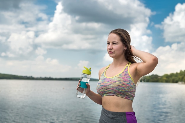 Hermosa deportista descansando junto al lago después del entrenamiento con una botella de agua en la mano