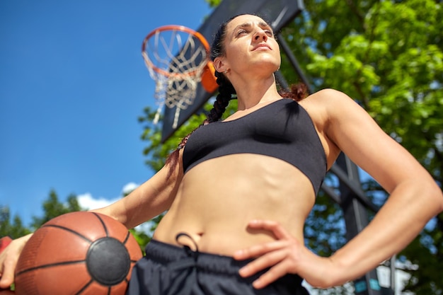 Hermosa y deportiva chica latina con una pelota de baloncesto debajo del aro en una cancha de baloncesto callejera. Motivación deportiva, estilo de vida saludable, espacio para copiar, banner publicitario.