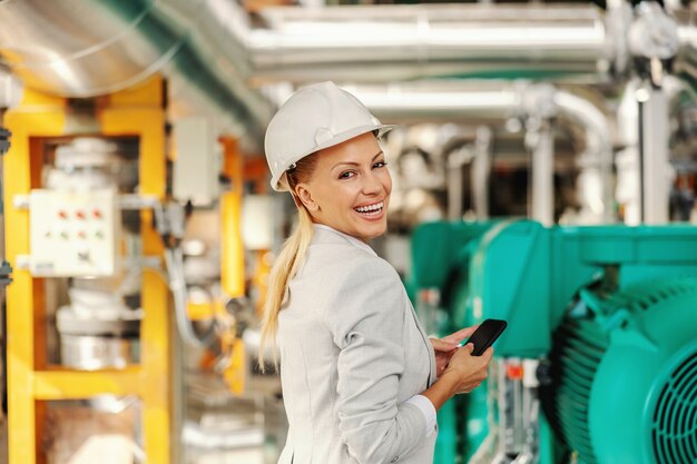 Hermosa directora ejecutiva rubia sonriente en traje con casco en la cabeza mirando por encima del hombro y mensaje de texto mientras está de pie en la planta de calefacción.