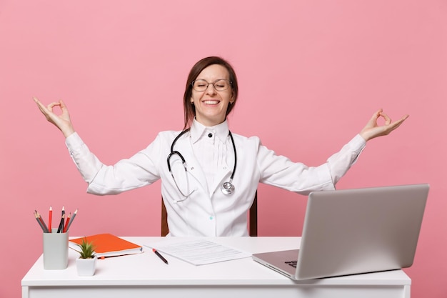 Hermosa doctora se sienta en el escritorio trabaja en computadora con documento médico en el hospital aislado sobre fondo de pared rosa pastel. Mujer con estetoscopio de gafas de bata médica. Concepto de medicina sanitaria