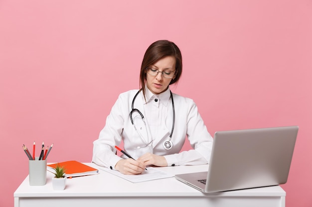 Hermosa doctora se sienta en el escritorio trabaja en computadora con documento médico en el hospital aislado sobre fondo de pared rosa pastel. Mujer con estetoscopio de gafas de bata médica. Concepto de medicina sanitaria