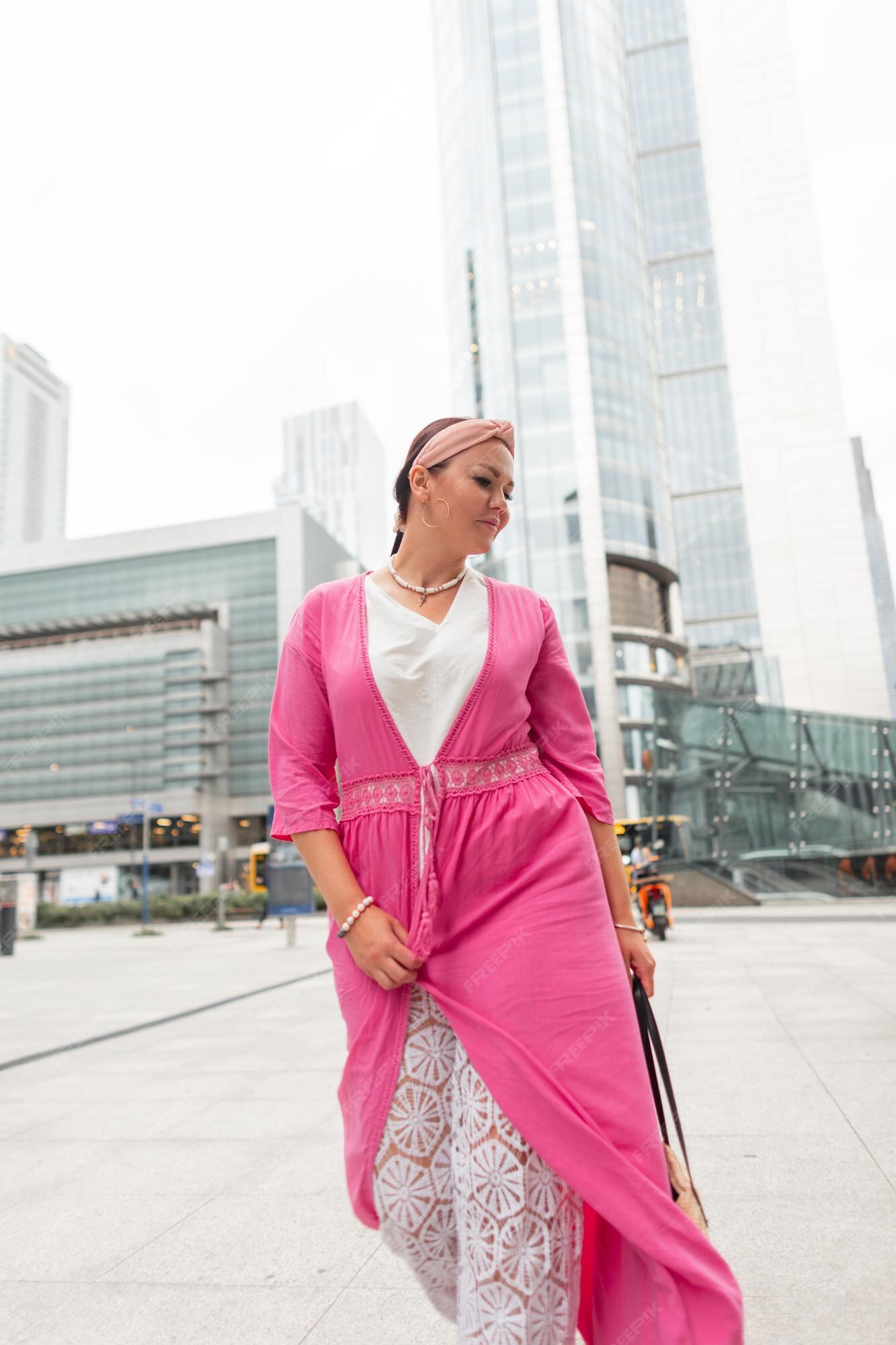 Hermosa y elegante chica de talla grande con ropa de playa de verano brillante de moda con un vestido rosa y pantalones de encaje blanco camina en ciudad moderna con edificios