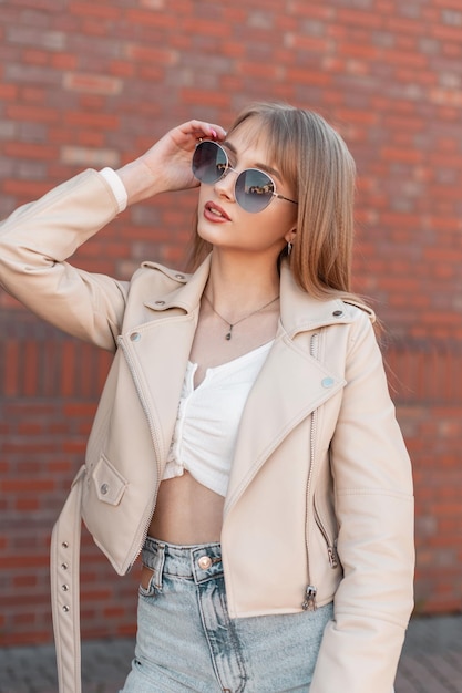 Hermosa y elegante chica zoomer con ropa de moda con una chaqueta blanca de cuero y una blusa con jeans vintage de pie cerca de una pared de ladrillos y con gafas de sol redondas