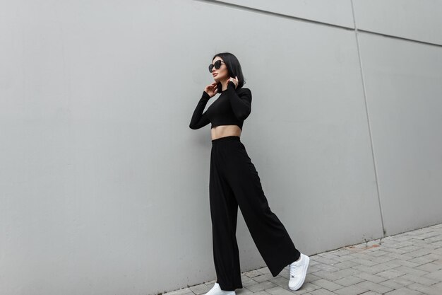 Hermosa y elegante modelo de moda con ropa negra de moda con gafas de sol de moda con zapatillas blancas caminando cerca de una pared gris en la ciudad