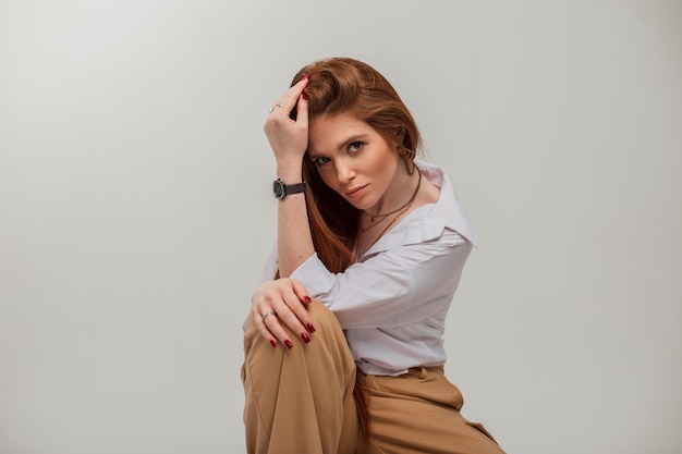 Hermosa y elegante mujer de negocios exitosa con cabello largo y rojo en ropa de moda con una camisa blanca sentada en una silla sobre un fondo blanco