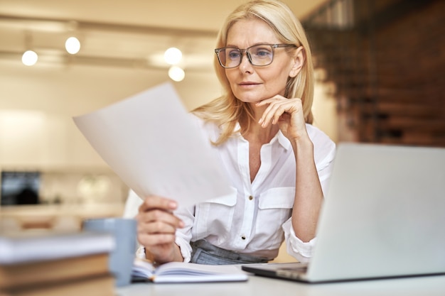 Hermosa empresaria madura leyendo documentos usando la computadora portátil mientras está sentada en su escritorio en la oficina