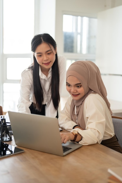 Una hermosa empresaria musulmana asiática usando su computadora portátil mientras habla de trabajo con su colega