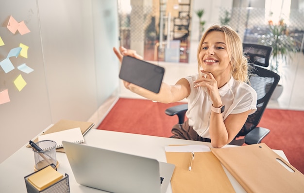 Hermosa empresaria tomando fotos con el teléfono inteligente y sonriendo mientras está sentado en la mesa de la oficina con el portátil