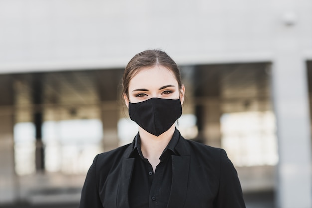 Hermosa empresaria en un traje negro en una máscara médica negra y guantes en la ciudad en cuarentena y aislamiento. Pandemia COVID-19. Enfoque selectivo