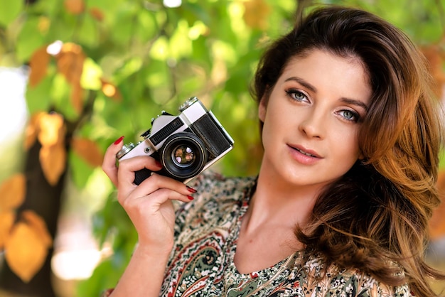 Hermosa y encantadora mujer sosteniendo una cámara SLR retro y tomando fotos en un parque de otoño en un día soleado