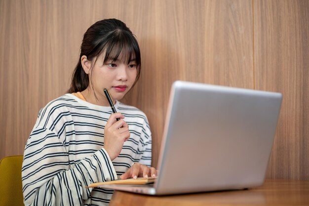 Una hermosa y enfocada mujer asiática está haciendo su tarea y usando su portátil en una mesa en una cafetería