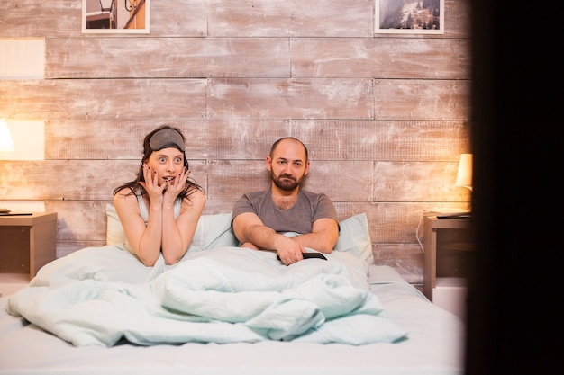 Hermosa esposa en pijama viendo una película impactante en la televisión junto con su marido aburrido.