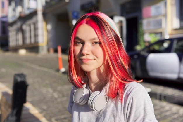 Hermosa estudiante adolescente feliz sonriente con auriculares de peinado de colores de moda, calle de fondo soleado de la ciudad de verano