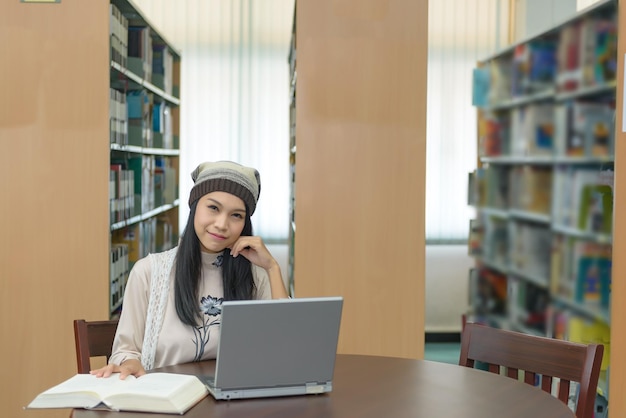 Hermosa estudiante asiática que usa una computadora portátil para estudiar en la biblioteca que lee antes de abrir la computadora portátil