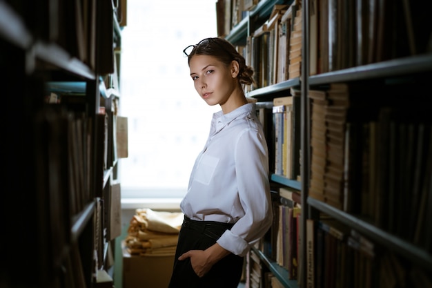 Hermosa estudiante en camisa se encuentra entre las filas de la biblioteca, estanterías de libros. Foto oscura